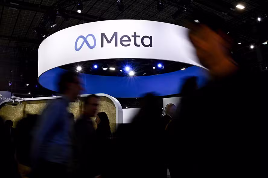 Estande da Meta em evento de tecnologia, com grande estrutura circular suspensa exibindo o logotipo e o nome da empresa. Visitantes circulam pelo espaço, aparecendo desfocados em primeiro plano, sob iluminação azul e branca.